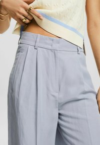 Kobieta w niebieskich plisowanych spodniach i kremowym bezrękawniku z lekkim niebieskim paskiem, ręką poprawiająca bluzkę w okolicy talii.