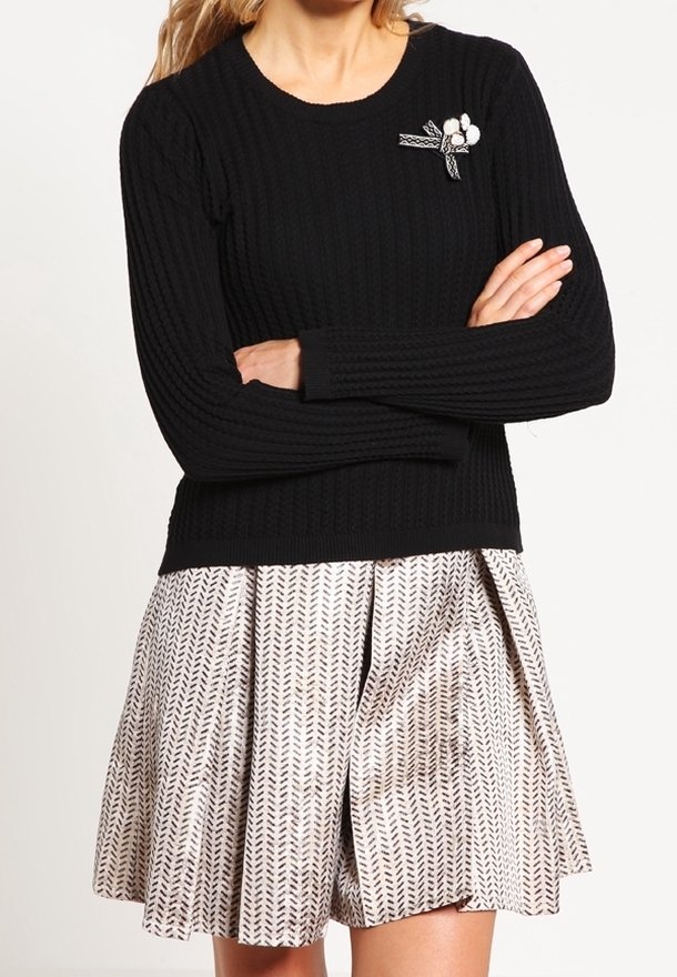 Femme portant un pull noir côtelé avec une broche fleurie et une jupe beige plissée à motifs, les bras croisés à la taille.