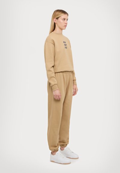 Hanorac bej cu text brodat, asociat cu pantaloni jogger bej asortați, amândouă dintr-o țesătură moale. Adidașii albi completează ținuta.
