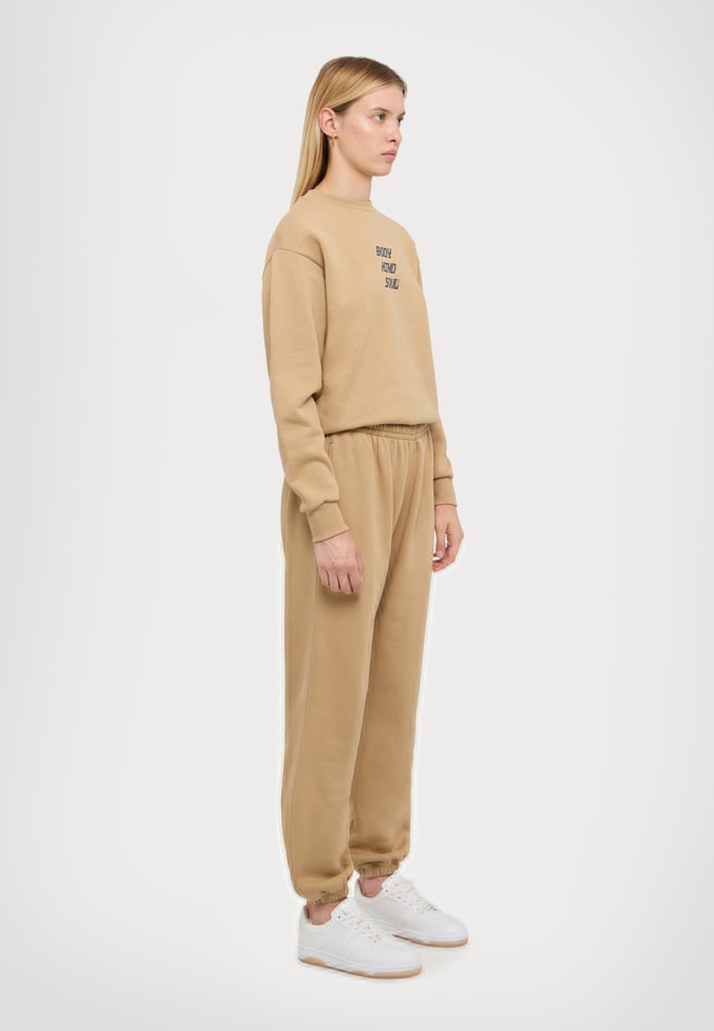 Beige sweatshirt met geborduurde tekst, gecombineerd met bijpassende beige joggingbroek, beide van een zachte stof. Witte sneakers maken de outfit af.