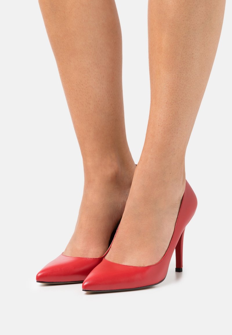 Jambes portant des escarpins rouges à bout pointu avec des talons aiguilles, se tenant devant un fond blanc.