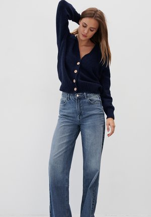 Granatowy dziany kardigan z dekoltem w serek, ozdobiony pięcioma dużymi beżowymi guzikami, połączony z jasnoniebieskimi spodniami o wysokim stanie i szerokich nogawkach.