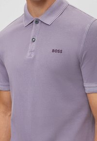 Polo a manica corta e aderente in viola chiaro con tessuto strutturato, due bottoni sulla patta e logo "BOSS" viola scuro sul petto.