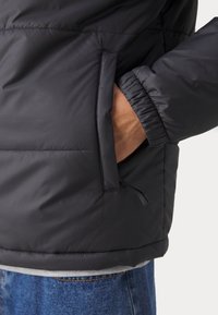 Main d'une personne dans la poche avant d'une veste matelassée noire avec poignets élastiques, portant un jean bleu et une chemise grise en dessous.