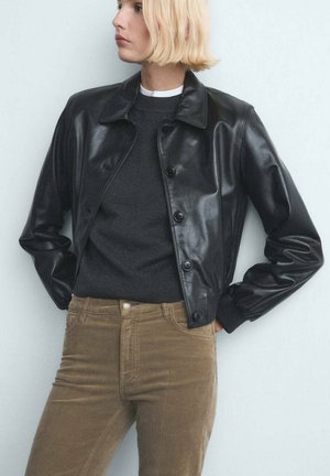 Blonde Person trägt eine schwarze Lederjacke mit Knöpfen über einem dunkelfarbenen Pullover und braunen Hosen, steht mit den Händen in den Taschen vor einer hellen Wand.