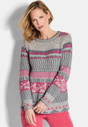 Eine Frau mit blonden Haaren trägt Ohrringe, einen grauen und rosa gemusterten Pullover mit langen Ärmeln und rosa Hosen und steht vor einem weißen Hintergrund.
