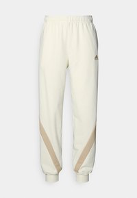 Beżowe spodnie dresowe z elastycznym pasem i mankietami, ozdobione beżowymi, skośnymi paskami na dolnych partiach nogawek oraz małym logo Adidas na górnej lewej stronie uda.