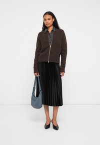 Γυναίκα που φορά καφέ φούτερ με φερμουάρ, μαύρη πλισέ μίντι φούστα, μαύρες γόβες, κρατώντας μπλε τσάντα ώμου, που στέκεται μπροστά σε λευκό φόντο.