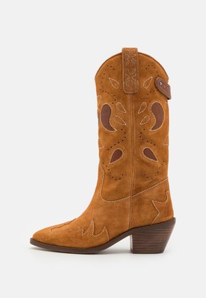 Ruskeat mokkanahkaiset cowboy-saappaat, joissa on terävät kärjet, koristeelliset leikkaukset ja valkoiset tikkaustyöt. Niissä on lyhyt pino korko ja vetolenkki.