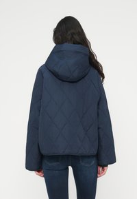 Μπλε ναυτικού μπουφάν με καπιτονέ ύφασμα και κουκούλα, με τετράγωνο σχήμα και υφή διαμαντένιου μοτίβου. Η πίσω όψη δείχνει φαρδιά μανίκια και δεν υπάρχουν εμφανή μεταλλικά εξαρτήματα.