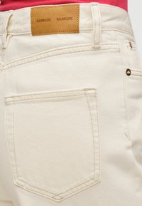 Jean en denim blanc avec une texture douce, doté d'une étiquette en cuir marron à la taille et d'un détail sur la poche arrière. Design simple et classique.