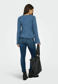 Frau in blauer Rippstrickjacke, dunklen Skinny-Jeans, schwarzen Stiefeletten mit Knöchelschaft, die eine große schwarze Ledertasche hält und vor einem weißen Hintergrund steht.