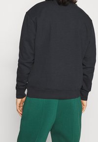 Sudadera negra, mezcla de algodón, cuello redondo, puños y dobladillo acanalados, corte holgado, costuras visibles, combinada con pantalones verdes.