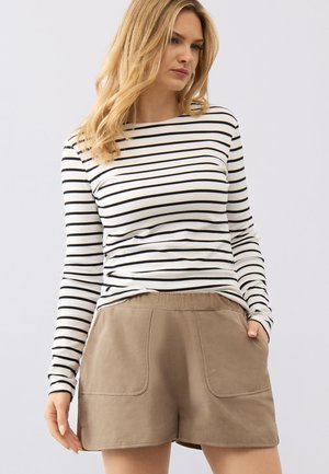 Kvinde iført en hvid langærmet skjorte med sorte vandrette striber og beige shorts med store forlommer, stående foran en hvid baggrund.