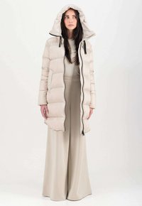 Piumino beige chiaro con cappuccio, caratterizzato da un design trapuntato, zip frontale e cinturini neri in evidenza. Abbinato a un outfit beige coordinato.