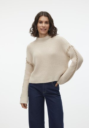 Beige strikkegenser med høy hals og løs passform. Har sidesnarer detaljer på ermene, brukt sammen med mørkeblå vide jeans.