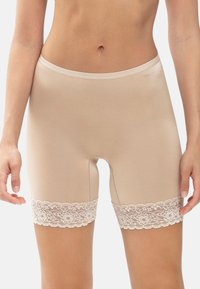 Beiga sömlösa damsorts med blommig spetsdetalj vid låren, bärs av en stående modell med händerna avkopplade längs sidorna.