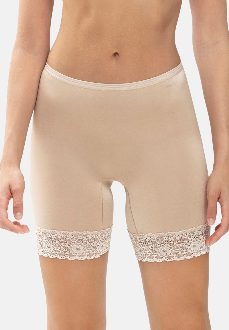 Beiga sömlösa damsorts med blommig spetsdetalj vid låren, bärs av en stående modell med händerna avkopplade längs sidorna.