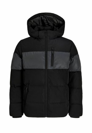 Garçon portant une doudoune noire, un pantalon camouflage, des baskets beiges, une chemise blanche et une casquette sombre, debout et souriant sur un fond blanc.