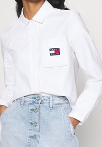 White button-up shirt with a chest pocket featuring a black, red, and blue logo. Paired with light blue denim skirt, silver button details.