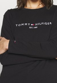 Maglietta a maniche lunghe nera con logo "TOMMY HILFIGER" bianco e testo "EST. 1985". Presenta un piccolo accento rosso e bianco sulla manica.