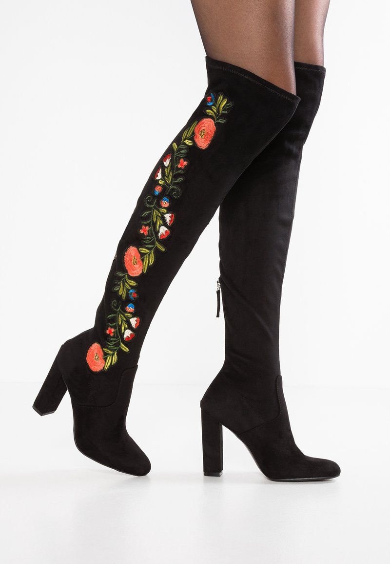 Bottes cuissardes en daim noir avec broderies florales en rouge, bleu et vert, dotées d'un talon bloc et d'une fermeture éclair latérale.