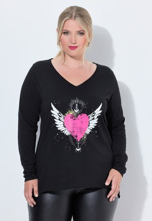 Frau mit blonden Haaren, die ein schwarzes Langarmshirt mit einem pinken Herz und weißen Flügeln auf der Vorderseite trägt.