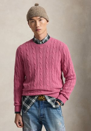 Man met een beige gebreide muts, een roze kabelgebreide trui over een groene geruite blouse en blauwe verweerde spijkerbroek met een decoratieve riemgesp, staand.