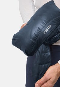 Marineblaues Pufferjackett, in eine kompakte Form verpackt, mit einem „STOW AWAY“-Etikett, glattem, glänzendem Stoff und minimalen Nähten.