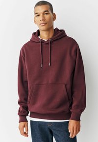 Burgunderfarbener Kapuzenpullover mit einer Fronttasche, verstellbaren Kordeln und Rippenbündchen am Ärmel und Saum, getragen über einem weißen Shirt.