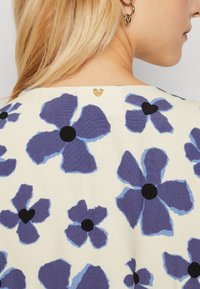 Tissu à motif floral avec de grandes fleurs violettes et bleues, dotées de centres noirs, sur un fond crème. Accent en cœur doré au niveau de l'encolure.