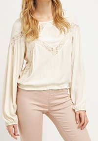 Krämfärgad blus med långa, samlade ärmar och spetsdetaljer längs halsen, ihop med ljusbeiga byxor. Slät tygtextur.