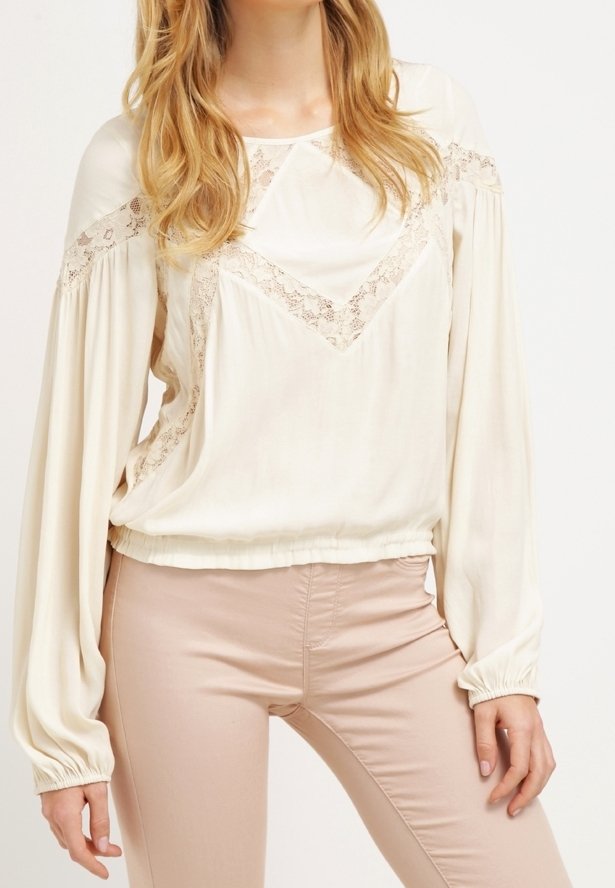 Krämfärgad blus med långa, samlade ärmar och spetsdetaljer längs halsen, ihop med ljusbeiga byxor. Slät tygtextur.