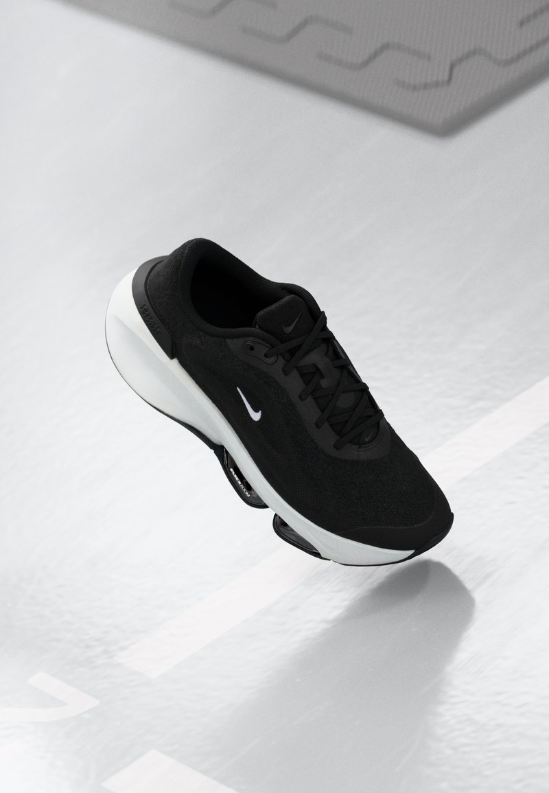 Chaussure de course Nike noire avec logo blanc et semelle blanche épaisse, suspendue au-dessus d'une surface blanche avec des ombres et des marquages au sol.