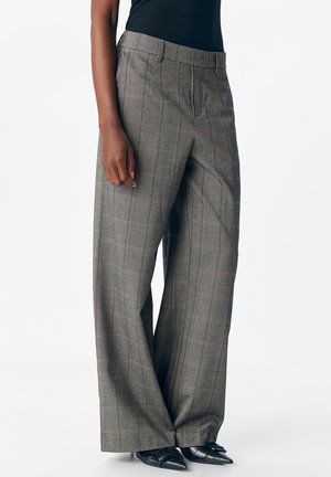 Femme portant un pantalon à larges jambes à carreaux gris, des chaussures noires à bout pointu et un haut noir sans manches sur un fond blanc.