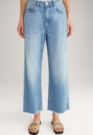 Persoon die lichtblauwe brede broekspijpen draagt met onafgewerkte zomen en beige slip-on sandalen met goudkleurige kettingdetail.