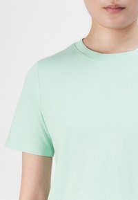 Záber zblízka osoby nosiacej jednofarebné svetlozelené tričko s krátkym rukávom a okrúhlym výstrihom na bielom pozadí.