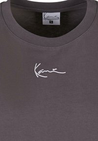 Harmaa puuvillapaita, jossa on pyöreä kaula-aukko. Vasemmalla puolella valkoinen brodeerattu signeeraus, joka näyttää sileältä. Koko S.