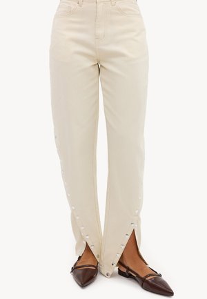 Pantalon couleur crème avec des boutons-pression le long des jambes extérieures, porté avec des ballerines marron à bouts pointus, visible de la taille aux pieds.