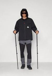 Črna jakna z zadrgo in kapuco, z dvema žepoma na prsih ter trikotnim poudarkom. Kombinirana s sivo hlačami in črnimi pohodnimi čevlji, drži pohodne palice.