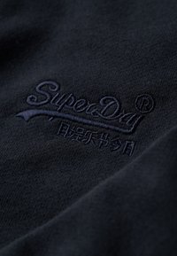 Mørkeblåt stof med et tekstureret, broderet logo "SuperDry" med dekorative elementer. Materialet fremstår blødt med en subtil glans.