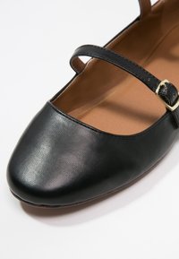 Chaussure Mary Jane en cuir noir avec un bout arrondi, une bride unique avec boucle et une texture lisse. Intérieur de couleur fauve contrastante.