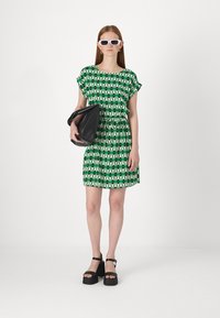 Abito verde con motivo triangolare, maniche corte, vita cintrata, abbinato a una borsa nera e sandali con piattaforma. Gli accessori includono occhiali da sole bianchi.