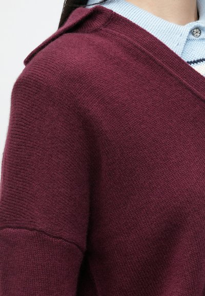 Burgundi kötött pulóver, amely textúrált felülettel, kapucnival és széles ujjakkal rendelkezik, világoskék galléros inggel, csíkos mintázattal a réteg alatt.