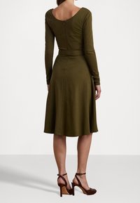 Olivová dlouhá šaty s fitted korzetem a rozšířenou sukní, s výstřihem na zádech a páskem v pase. Nosené s hnědými lodičkami na vysokém podpatku.