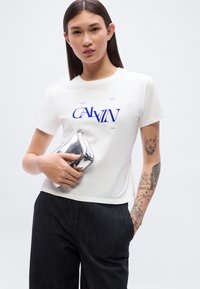 Λευκό βαμβακερό μπλουζάκι με μπλε τύπωμα "CALVIN KLEIN", κοντά μανίκια, συνδυασμένο με μαύρο υφασμάτινο παντελόνι και ασημένια τσάντα χειρός.