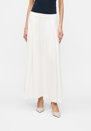 Donna che indossa una lunga gonna bianca con coulisse in vita, top nero senza maniche e tacchi beige con punta affilata, in piedi davanti a uno sfondo bianco.