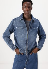 Denim jacket featuring a front zipper, two chest pockets, and a pattern of light blue shapes on a darker blue background.