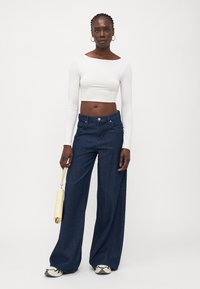 Top corto de manga larga blanco combinado con vaqueros de denim oscuros de pierna ancha. La modelo sostiene un bolso amarillo claro y lleva zapatillas blancas y negras.