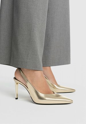 Złote metaliczne sandały na obcasie typu slingback z cienkim obcasem szpilkowym i szpiczastym noskiem, zestawione z szarymi szerokimi spodniami o gładkiej fakturze.